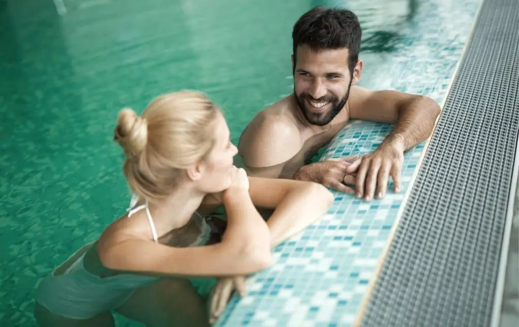 terme salsa Moški s temnimi lasmi in brado se toplo smehlja svetlolasi ženski z lasmi spetimi v figo, medtem ko oba počivata na robu termalnega bazena. Ženska v belem kopalnem kostumu se naslanja na rob, obkrožena s turkizno vodo v mirnem okolju term "terme salsą".